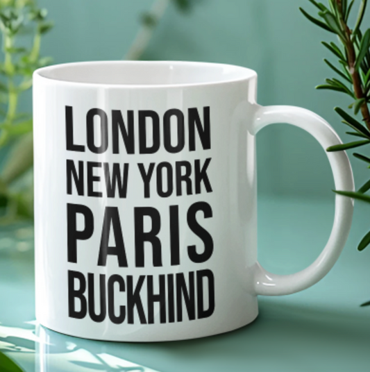 Buckhind Mug in white with bold black city name lettering, on a pale green background beside greenery