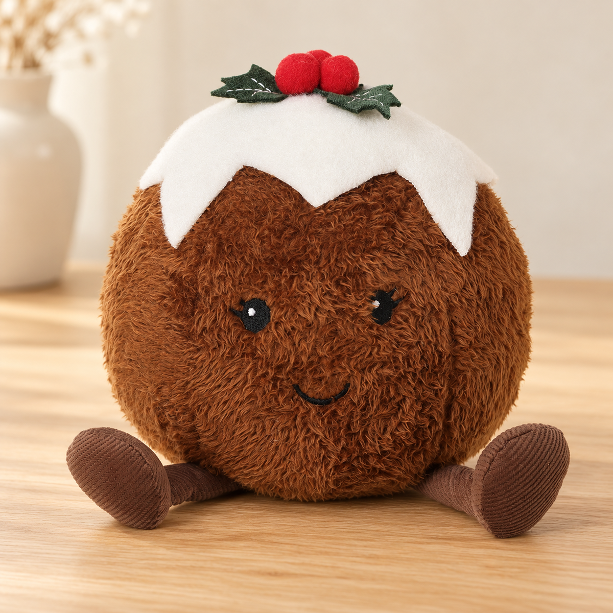A round brown plush pudding character toy with a white felt hat, red berries, green holly leaves, and small brown corduroy legs, sitting on a light wooden surface.