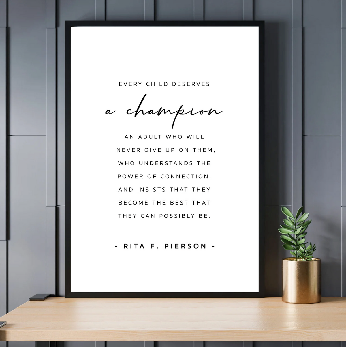 A Champion - Teacher print in a black frame on a wooden desk with a small plant against a dark wall