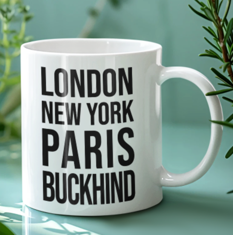 Buckhind Mug in white with bold black city name lettering, on a pale green background beside greenery