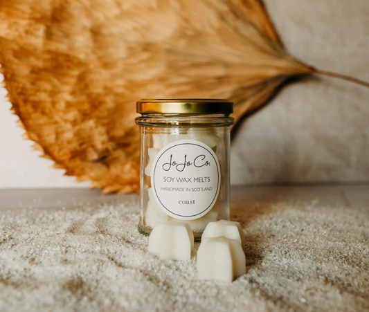 JoJo Co. Coast Soy Luxury Wax Melts, glass jar and loose white melts on sand with a dried palm leaf
