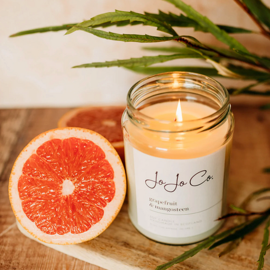 JoJo Co. Grapefruit & Mangosteen Luxury Candle in a clear glass jar beside a halved grapefruit and green leaves