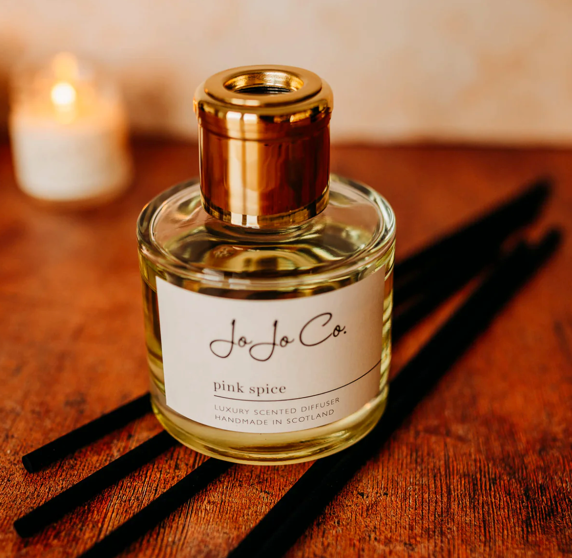 JoJo Co. Pink Spice Luxury Diffuser, a clear glass bottle with gold cap and cream label, on a wooden surface