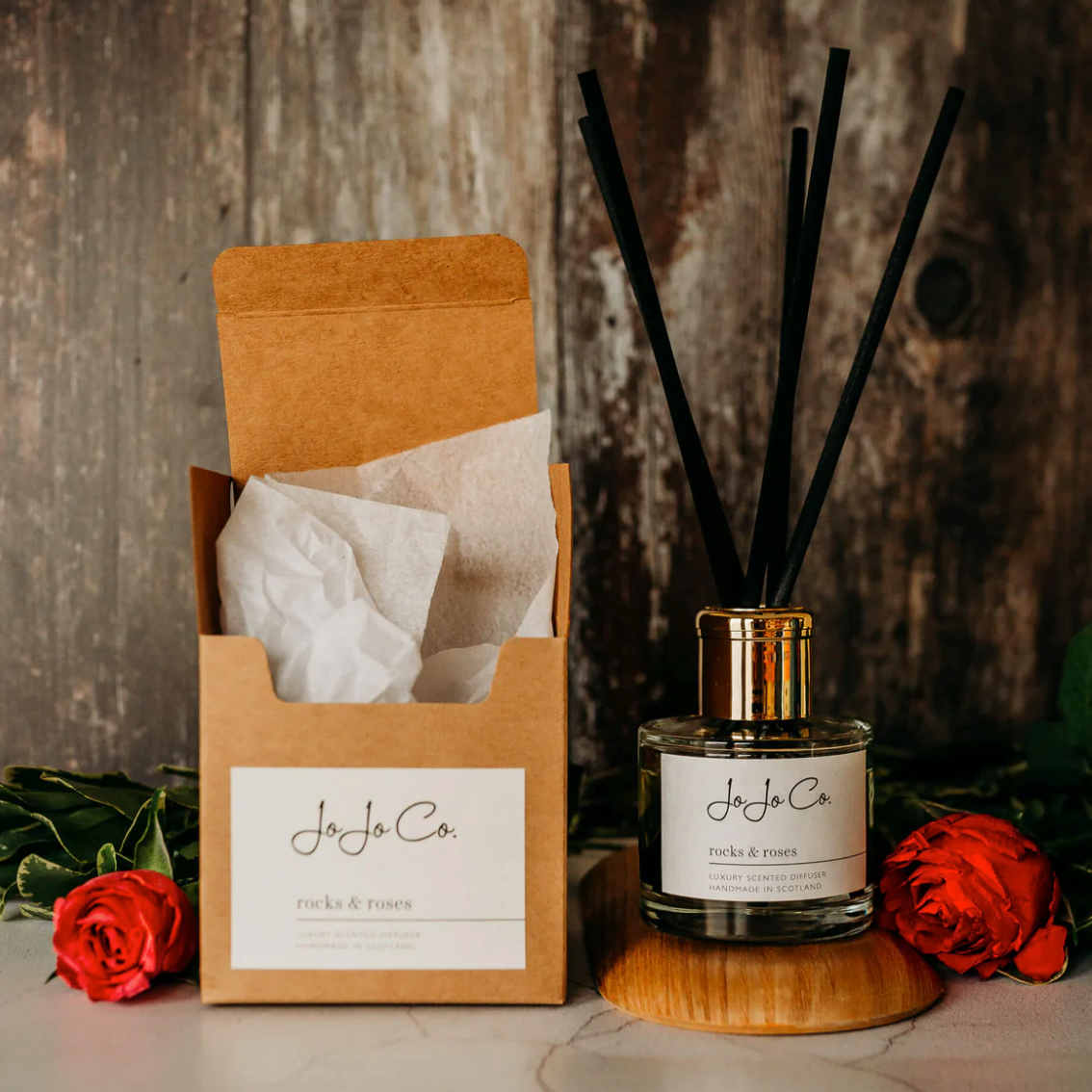 JoJo Co. Rocks & Roses Luxury Diffuser with black reeds, beside its kraft box and red roses on a wood board