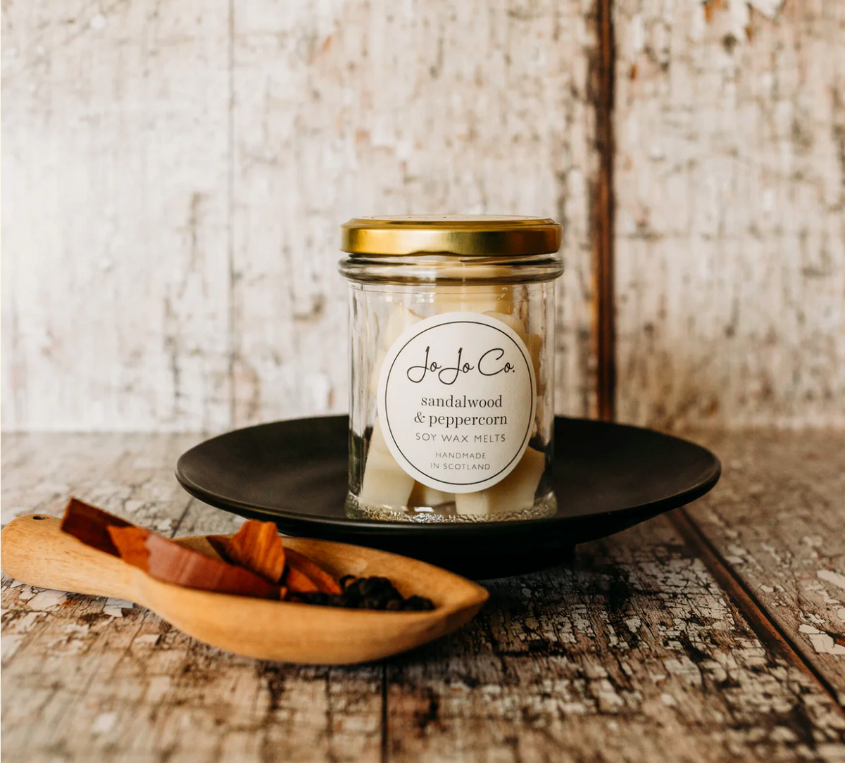 JoJo Co. Sandalwood & Peppercorn Soy Luxury Wax Melts in a glass jar with gold lid, on a rustic wooden table