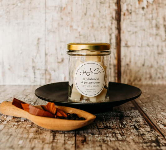 JoJo Co. Sandalwood & Peppercorn Soy Luxury Wax Melts in a glass jar with gold lid, on a rustic wooden table