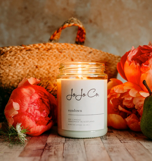 JoJo Co. Sundown Luxury Candle in a glass jar with cream label, among red flowers and a woven basket