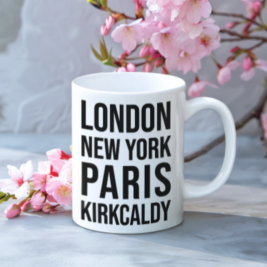 Kirkcaldy Mug, a white mug with bold black London New York Paris Kirkcaldy text, beside pink blossom