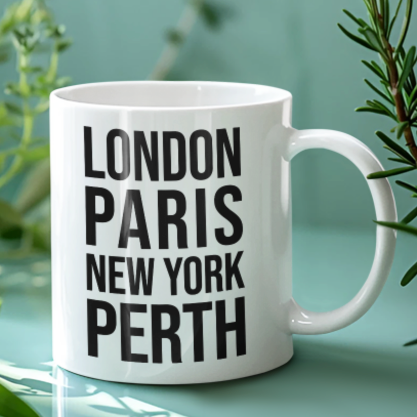 Perth Mug in white with black city name lettering, on a pale green surface beside greenery