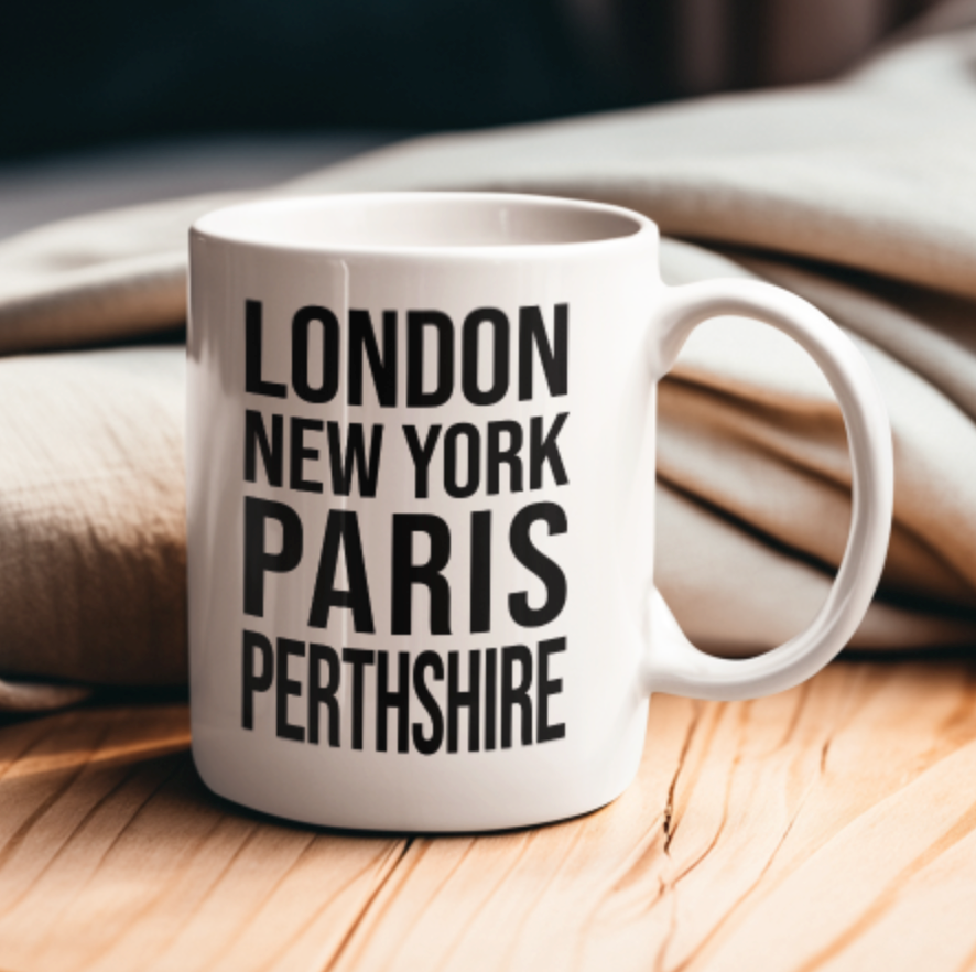 PERTHSHIRE Mug, a white mug with London New York Paris Perthshire text, on a wooden table with a knit throw