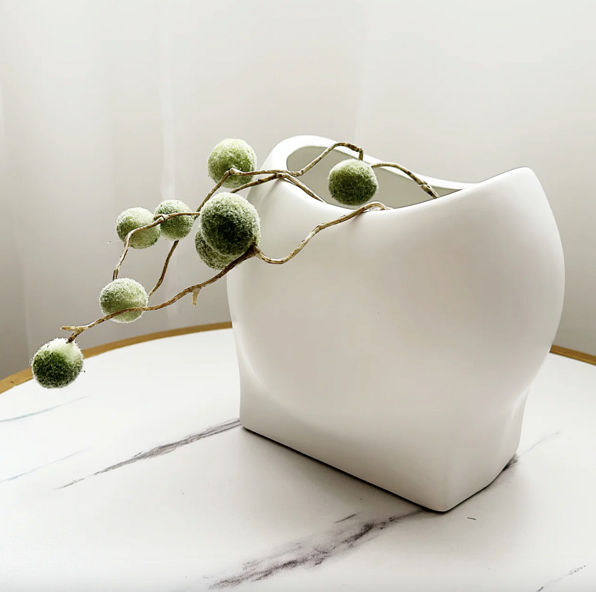 Poppy Vase - White, a matt white sculptural vase with a curved rim, holding trailing greenery on marble