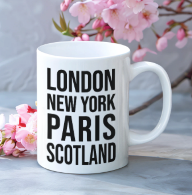 Scotland Mug in white with bold black city lettering, beside a sprig of pink blossom on a marble surface