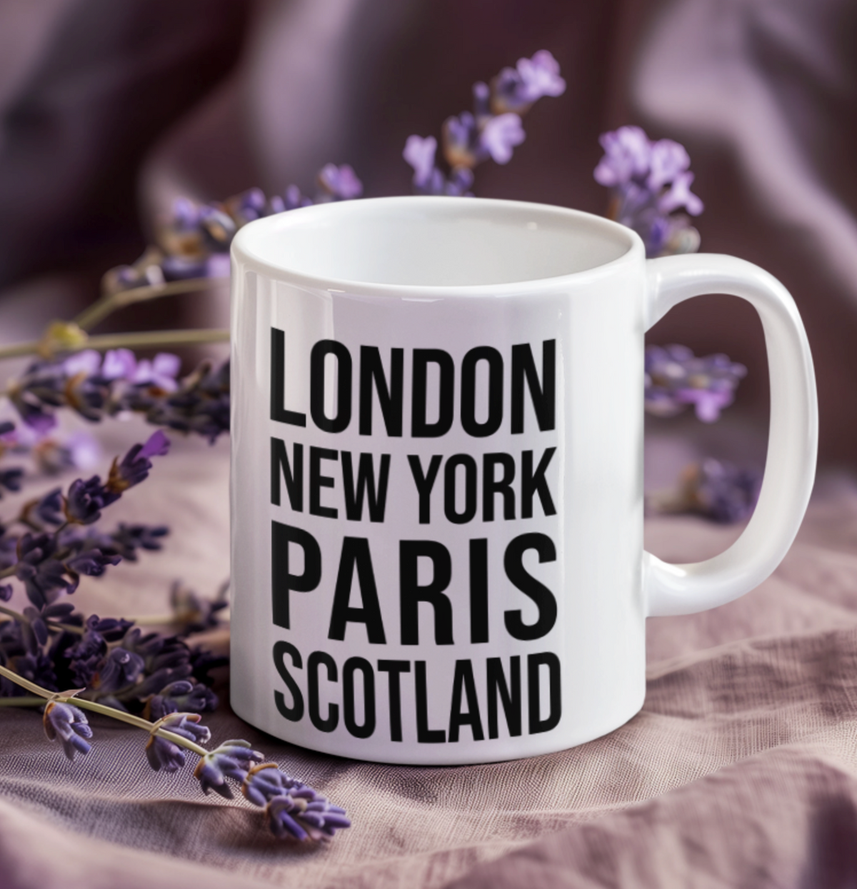 Scotland Mug, a white mug with black London New York Paris Scotland text, on purple fabric with lavender